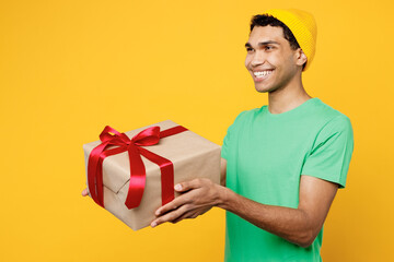 Wall Mural - Side view young man of African American ethnicity wears casual clothes green t-shirt hat hold in hand present box with gift ribbon bow look aside isolated on plain yellow background Lifestyle concept.