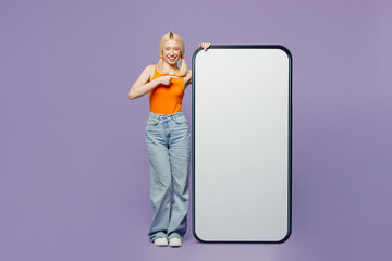 Wall Mural - Full body young blonde woman wear orange tank shirt casual clothes point index finger on big huge blank screen mobile cell phone smartphone with area isolated on plain pastel light purple background.