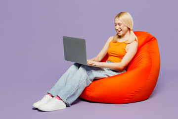 Full body young blonde IT woman wear orange tank shirt casual clothes sit in bag chair hold use work on laptop pc computer isolated on plain pastel light purple background studio Lifestyle concept.