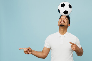 Wall Mural - Young fun cool man fan wearing basic t-shirt cheer up support football sport team holding in hand soccer ball on head point aside watch tv live stream isolated on plain pastel blue color background.