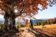© Olha - Family is hiking in mountains. Children with backpacks stand near large lonely tree with autumn leaves.