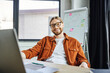 © LIGHTFIELD STUDIOS - happy and optimistic bearded businessman in eyeglasses and trendy shirt sitting at laptop and looking at camera next to flip chart on blurred background in contemporary office