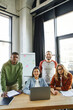 © LIGHTFIELD STUDIOS - diverse group of optimistic, young and multicultural business people looking at camera near laptop, documents and flip chart with graphs on background in modern office space, creative collaboration