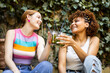 Two cute multiracial girls are happily toasting with some flavored cocktails in a place with vegetation. Concept of multiethnic couple drinking mojitos, toast with combined.