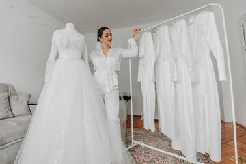 Wall Mural - a beautiful bride poses in a white robe next to her wedding dress and her friends' dresses on a hanger