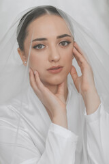 Wall Mural - Portrait of a beautiful bride girl with stylish hairstyle and makeup in a white robe, under a veil, gently holding her face with her fingers