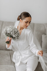 Wall Mural - Portrait of a beautiful bride girl with stylish hair and makeup in a white robe sitting on a sofa in her room with a bouquet of gypsophila flowers and holding her veil