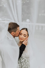 A stylish couple of European newlyweds. Smiling bride in a white dress. The groom, dressed in a classic black suit, white shirt, kisses the bride on the temple under the veil. Wedding in nature