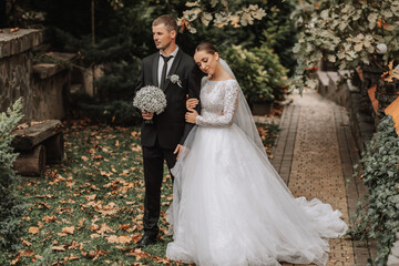 Wall Mural - A stylish couple of European newlyweds. A smiling bride in a white dress leaned against the groom's shoulder. Groom dressed in a classic black suit. Wedding in nature