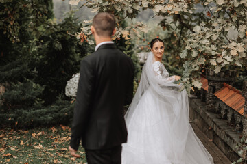 Wall Mural - A stylish couple of European newlyweds. A smiling bride in a white dress looks at the groom and flirts with him. Groom dressed in a classic black suit. Wedding in nature