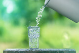 Fototapeta Łazienka - Water flows into a glass placed on a wooden bar.Water splash in glass Select focus blurred background.Drink water pouring in to glass over sunlight and natural green background.Nature conservation . 