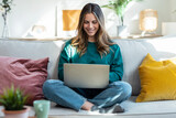 Fototapeta Lawenda - Beautiful kind woman working with laptop while sitting on couch in living room at home