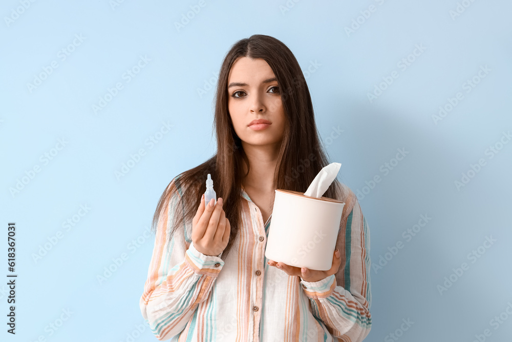 Allergic young woman with nasal drops and tissue box on blue background