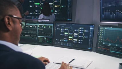 IT technical specialist sits in front of computer with blockchain network database in modern monitoring office. Team of software programmers work with real-time analysis charts on big digital screens.