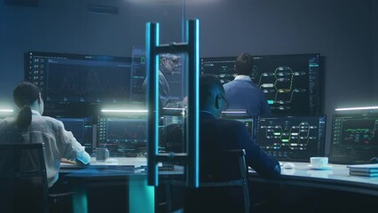 Multiethnic team of IT technical specialists and software programmers work with data server, analysis charts and blockchain network on computers and big digital screens in monitoring office at night.