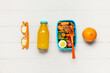 Bottle of juice, eyeglasses, orange and lunchbox with tasty food on white wooden background