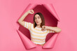 Beautiful caucasian brunette woman wearing striped t shirt posing in torn pink paper wall standing with raised arms surprised face pout lips looking directly at camera.
