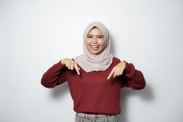 Wall Mural - Portrait of Asian Muslim woman smiling pointing fingers down, inviting customers to special event isolated over white background