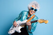 © wolfhound911 - Portrait of a cool trendy old woman playing a guitar on blue studio background. Generative AI