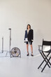 © LIGHTFIELD STUDIOS - fashion and style, studio photography, young asian woman in stylish look posing on grey background, blazer, white shirt and latex shorts, standing near electric fan, personal style, full length