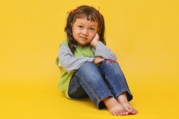 Wall Mural - Full body image of cute Asian baby girl sitting with hand on cheek isolated on yellow background