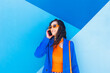 © VISTA by Westend61 - Happy fashionable woman wearing sunglasses talking on smart phone in front of blue wall