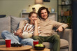© Seventyfour - Portrait of happy young couple watching movies at home and eating popcorn sitting on couch together