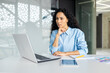 © Liubomir - Serious thinking woman working inside office at workplace with laptop, confident hispanic woman looking at technical task, brainstorming solution, business woman with curly hair looking at laptop