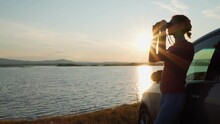 A Free Girl Driver Stands Next To The Car, Enjoying The View Of The Lake At Sunset. A Girl Traveler Looks Through Binoculars At A Beautiful Landscape And Sunset. A Beautiful Girl Travels Alone By Car.