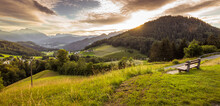 Wooden Resting Bank On The Slope Of A Hill In The Foreground, Overlooking The Valley Below In Berchtesgaden, Germany. It Is Evening, During Golden Hour, Clouds Are Covering The Sun. Summer, Day.