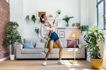 Wall Mural - Funny motivated woman dancing and listening to music with headphones while sweeping the house