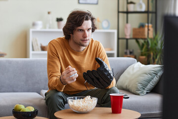 Portrait of emotional young man wearing baseball glove watching sports match on TV at home, copy space