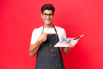 Wall Mural - Young artist caucasian man holding a palette isolated on red background pointing to the side to present a product