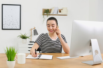 Canvas Print - Home workplace. Happy woman with pen and notebook talking on smartphone at wooden desk in room