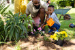 African american father looking at cute son watering fresh flowers with can in backyard