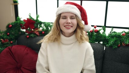 Sticker - Young blonde woman wearing christmas hat sitting on sofa at home