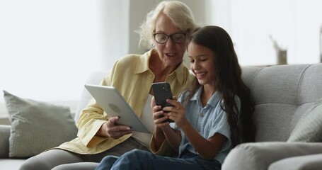 Little girl grandchild helps with modern digital tablet usage to aged granny sit on sofa with smart phone spend time on internet. Generations and wireless technology overuse, new application, software