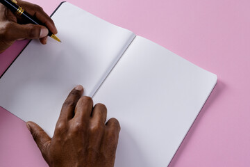 Poster - Hands of biracial man holding pen and writing in notebook with copy space on pink background