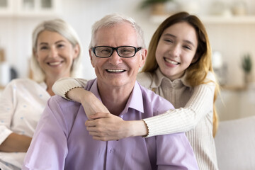 Wall Mural - Happy positive elderly grandfather posing at camera with hugging teenage grandkid girl and mature wife in background, enjoying leisure with family, good health, wellbeing