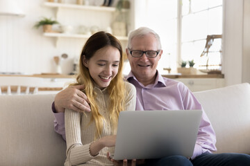 Wall Mural - Happy teenager kid and elder grandfather enjoying modern internet technology at home together, girl using application on laptop computer, teaching granddad to use online service