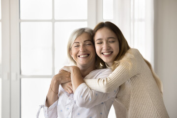 Wall Mural - Joyful positive teenage girl tightening beloved grandma with closed eyes and toothy smile. Granddaughter and grandmother enjoying close family relationship, hugging with love, tenderness