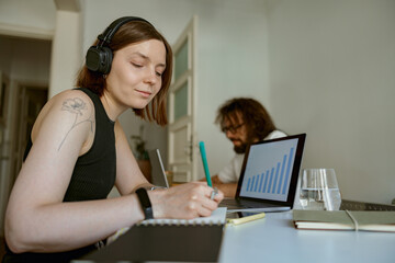 Wall Mural - Woman in headphones making notes on husband background while they working from home office