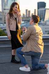 Wall Mural - Handsome man is doing surprise marriage proposal to his beautiful girlfriend on rooftop standing on his knee. Happy loving couple on romantic date on Saint Valentine's Day. Urban cityscape background