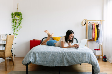 Happy young woman using laptop lying on bed at home. Millennial teenage girl working or studying online on computer in bedroom. Technology and domestic life concept.