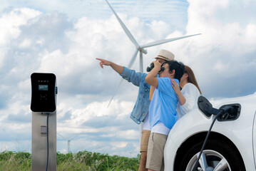 Concept of progressive happy family enjoying their time at wind farm with electric vehicle. Electric vehicle driven by clean renewable energy from wind turbine generator for charging station.