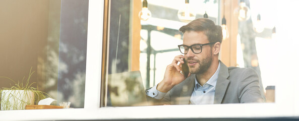 Wall Mural - young handsome businessman wearing eyeglasses talking by phone while working remotely in cafe