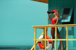 Lifeguard woman in red bikini and visor with binocular and lifebuoy on watch tower against turquoise sky