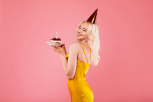 Positive Blonde Woman In Dress And Party Hat Holding Piece Of Cake With Candle And Smiling At Camera, Pink Background
