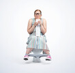 © afxhome - Funny young woman in glasses and a colored dress, eyes closed, sitting on the toilet, pushing, holding toilet paper in her hands, isolated on a light blue background