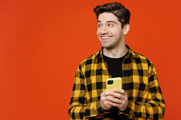 Wall Mural - Young smiling happy fun cheerful man wear yellow checkered shirt black t-shirt hold in hand use mobile cell phone look aside isolated on plain red orange background studio portrait. Lifestyle concept.